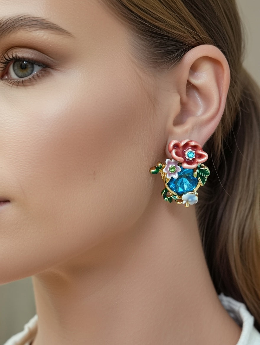 Close-up of floral enamel stud earrings. The vibrant blue central stone is framed by textured pink and white petals and emerald green leaves, creating a colorful, botanical aesthetic.
