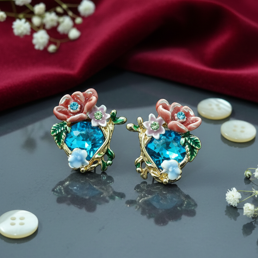 Hand-held pair of ornate floral earrings. Each piece centers on a heart-shaped turquoise crystal, accented by miniature enamel blossoms with rhinestone centers and delicate green foliage